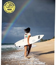 Person walking along the beach carrying a white Vans-branded surfboard with a rainbow in the background.